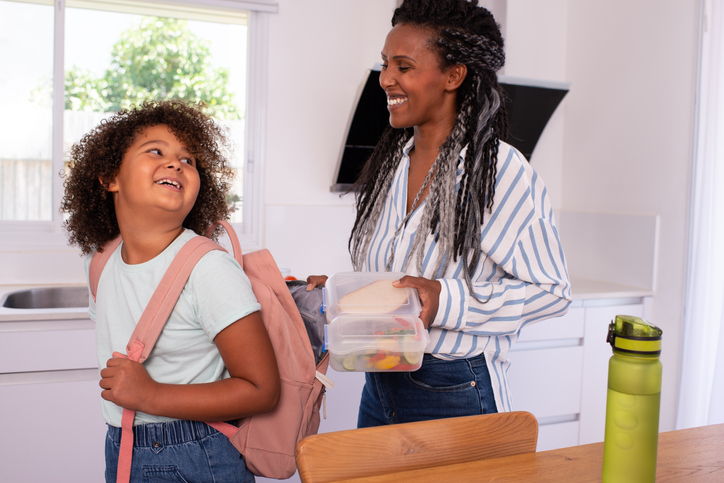 A mother helps her daughter to prepare for school, putting her lunch box and water bottle into a backpack.